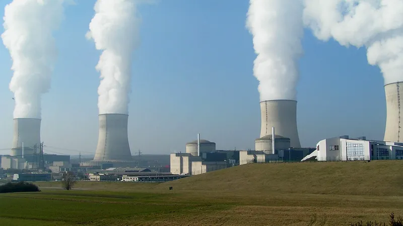 A imagem mostra uma vista panorâmica de uma usina nuclear com quatro grandes torres de resfriamento que emitem vapor de água contra um céu claro. O complexo inclui vários edifícios e estruturas industriais, com um campo gramado em primeiro plano.